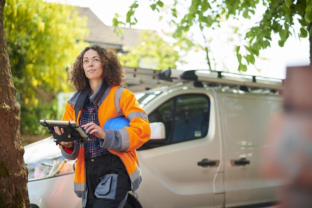 A woman holding an electronic tablet stands in front of a white van and looks up at something offscreen. She wears a bright orange high-visibility jacket and holds a blue hard hat under one arm. The van has an extendable ladder strapped to its roof.