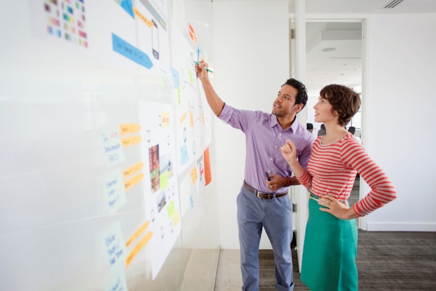 Two office employees, a man and a woman, stand before a whiteboard. The man is writing something on the whiteboard while the woman watches him.
