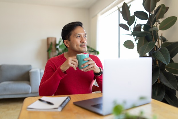 A man in a red sweater sits at a table with a mug in his hands and stares off wistfully into the distance. On the table in front of him are an open laptop and a notebook and pen. A leafy plant stands next to him; his gaze falls in its direction, although it doesn't seem like he's looking directly at it.