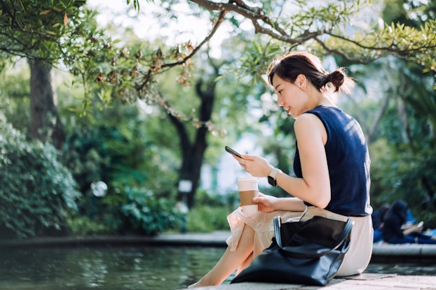  Woman sitting by a pond holding a coffee and scrolling through her phone.