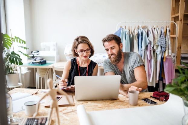 Two people sit at a wooden worktable and look at the open laptop in front of them. The person on the left is a woman with bobbed brown hair; she wears glasses and a red beaded necklace, and she is writing something down on a pad of paper. The person on the right is a man with a thick brown beard; he is wearing a gray T-shirt and pointing at something on the laptop screen. Also on the worktable are a couple of coffee mugs, a large roll of paper, a couple of notebooks, and a poseable wooden drawing model. In the blank-walled room behind the two people at the table are a rack of pastel clothing and a sewing machine on a small table.