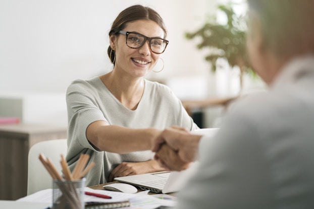 A woman wearing glasses sits at a desk and shakes hands with a person across from her, partially out of frame and out of focus.
