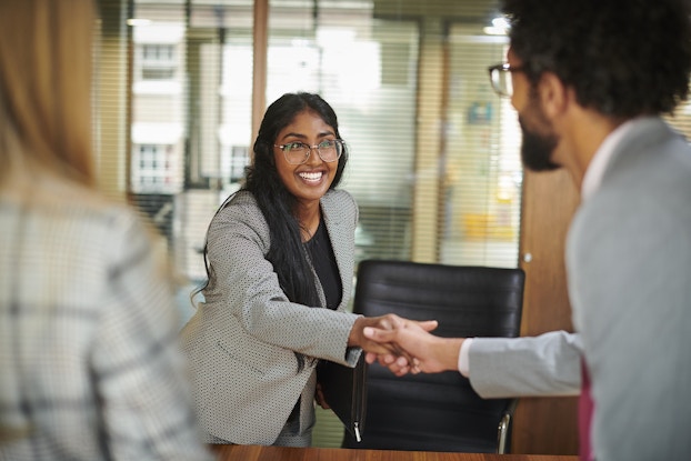 A young woman leans forward and shakes the hand of one of the two people standing in the foreground. The woman wears round glasses and a gray blazer; she has a large smile on her face. The man shaking hands with the woman has dark hair and a beard; he faces mostly away from the viewer. The second person in the foreground is out of focus and has long blonde hair and wears a white-and-black plaid shirt. Behind the young woman is a large window looking out at an office hallway.