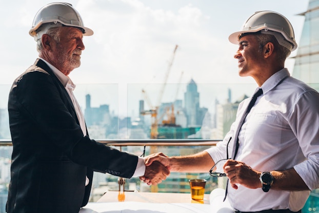  Two men in white hard hats shake hands. Behind them is a city skyline with a bright yellow crane working on one of the buildings.