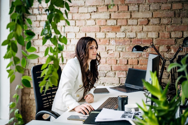 Woman working at a desk on a laptop in a modern-concept office with a red brick wall and greenery.