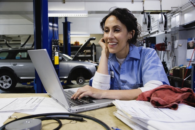 A mechanic sits at a table in her work garage, looking at the screen of an open laptop. She has one elbow resting on the table with her hand next to her smiling face, and she uses the other hand to type something on the laptop. The mechanic wears a light blue polo shirt over a white long-sleeved shirt, and her dark hair is tied back in a bun. In the background, a silver four-door truck is sitting with its hood open and its front tires missing.