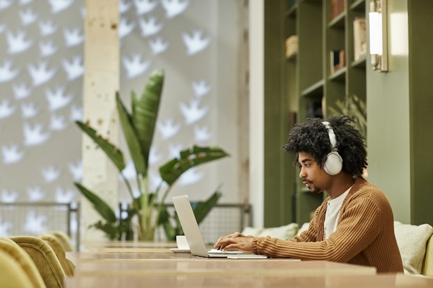 Man working on a laptop at a long table inside a coworking space wearing headphones.