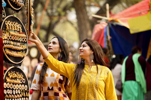 Two women with long dark hair look at hanging displays of large handmade earrings, most of them dangling and gold or copper. The earrings are stuck in circular boards hanging from an unseen structure. In the background are trees and part of a market stall around which other patrons clusters.