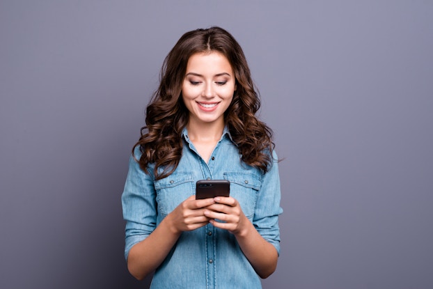 Woman standing looking at phone in front of a lilac-colored background.