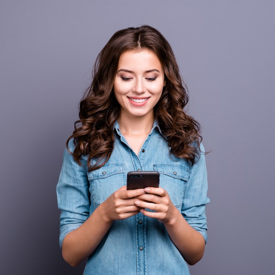 Woman standing looking at phone in front of a lilac-colored background.