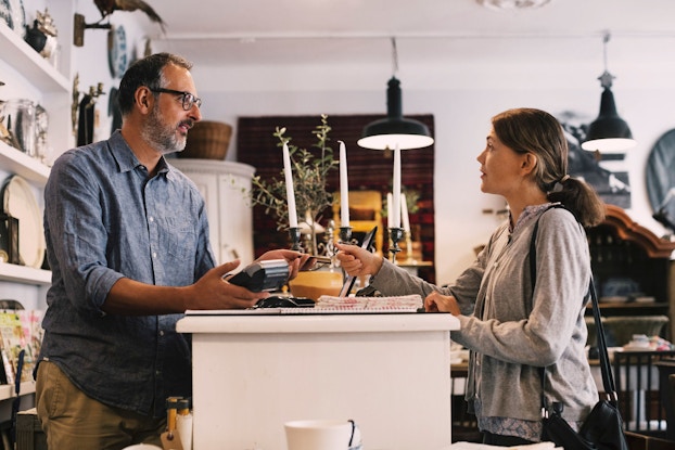  The owner of a small business home goods store hands a credit card back to a female customer. Both the owner and the customer are standing at the shop's counter.