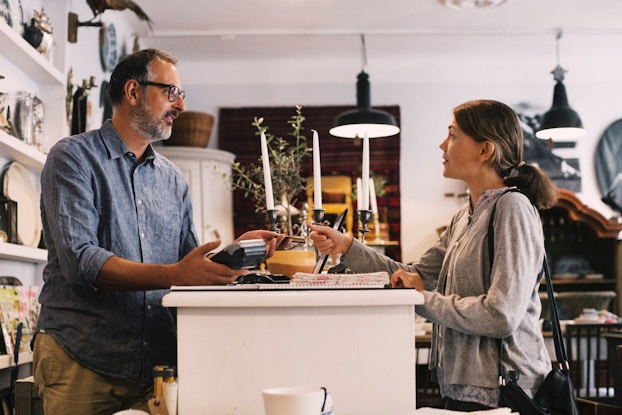 The owner of a small business home goods store hands a credit card back to a female customer. Both the owner and the customer are standing at the shop's counter.