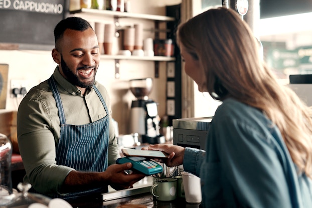 A man stands behind the counter of a cafe, accepting a mobile payment from a woman with a smile. The man has buzzed short hair and a beard, and he wears a navy blue apron over an olive green button-up shirt. He holds a teal-colored handheld payment system out for the woman to tap with her smartphone. The woman is facing away from the viewer; she has long brown hair and wears a dark blue jacket.