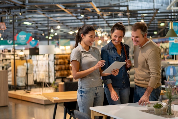  Three smiling people stand in a hardware/home improvement store. The woman on the left holds a tablet, which the man and woman on the right examine..