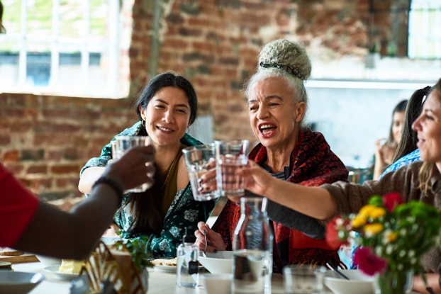  A group of people sit at a table and hold glasses of water outward for a toast. The two people in the center of the short are women, one wearing an emerald green patterned top and the other in a red top with her silver hair pulled into a bun on top of her head. The table in front of them is set with empty bowls, a glass water carafe, and a vase filled with flowers.