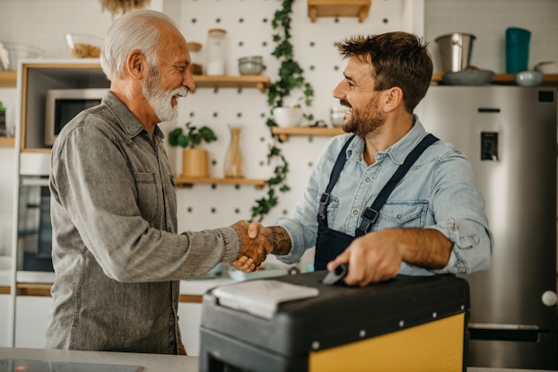 Two smiling men shake hands in a kitchen. The man on the left is taller and elderly, with thinning white hair and a white beard. He is wearing a long-sleeved gray collared shirt. The man on the right is using his other hand to grasp the handle of a large toolbox. He has dark hair and a dark beard, and he wears dark blue overalls over a chambray button-up shirt. In the background is a stainless steel refrigerator, several white cabinets, and a wall holding a few small wooden shelves.