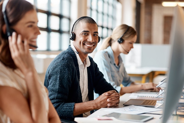  Three coworkers working in customer service, sitting at a desk wearing headphones.