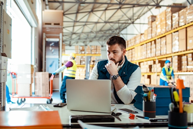 A man stands at a counter and looks at the screen of an open laptop. The man has dark hair and a beard and holds one hand to his chin as if in deep thought. Behind him is a large warehouse with yellow shelving units filled with cardboard boxes. To the left, two people in yellow high-visibility vests push and pull an orange dolly cart holding two piles of cardboard boxes into the warehouse.