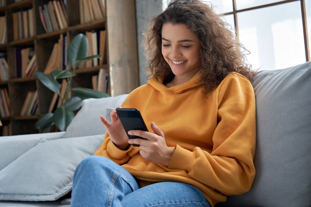 A woman in a baggy yellow sweater sits on a couch and looks down at the smartphone in her hands with a smile.