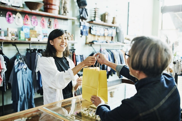 A young woman in a clothing store hands a yellow paper bag across a glass-topped counter to another, older woman. The wall behind the first woman is lined with shelves of boxed merchandise and racks of clothing.