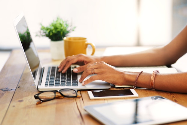  person's hands working on laptop beside a notebook, reading glasses, and a phone