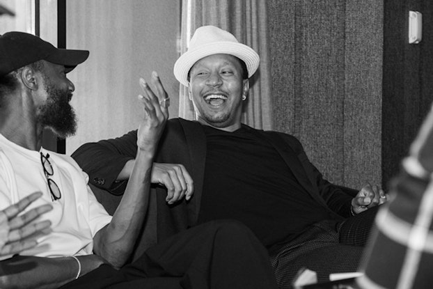 A black-and-white photo of Darius Davie, a Black man in a white fedora and dark shirt and jacket, sitting casually with a group of people. Davie is looking to his right and laughing at something said by the man next to him.