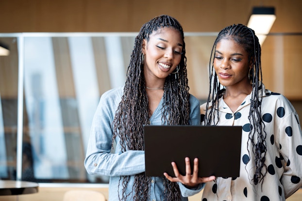 Two young women discuss a marketing project in an office. One of the women is holding a laptop computer in her right hand. Both women are looking at the computer.