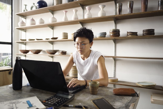  A young woman sits at a table in a workshop typing something on a laptop. The woman has a serious look on her face; she has short dark hair and wears a white T-shirt and glasses. Behind her are shelves holding ceramic bowls and vases.