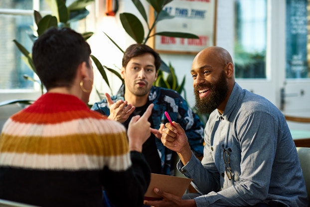 Three people sit in a group and talk. The person sitting on the right is a smiling man with a shaved head and a thick beard; he is holding a red pen in one hand. The person sitting in the middle is a man with dark hair and stubble; he is speaking and gesturing at the person across from him with one hand. The person across from him, seated on the left side of photo, is facing away from the viewer and emphasizing their point by pointing in the air with one finger. This person has short dark hair and is wearing a striped sweater and gold hoop earrings.