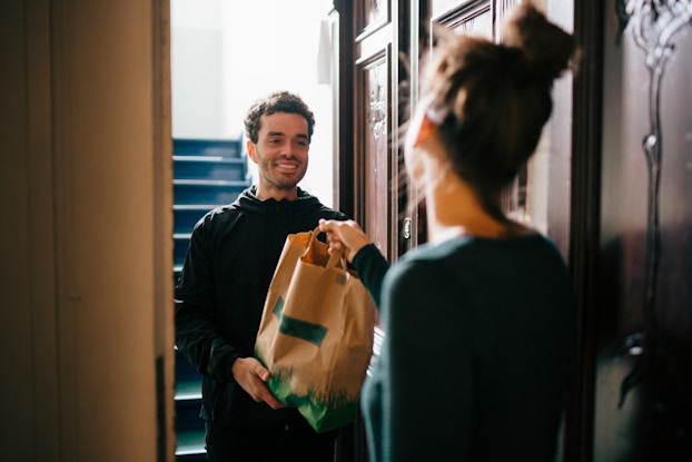 A man stands in a stairwell, seen through an open doorway. He holds out a paper bag with a smile. The man is wearing a black hoodie; he has short curly hair and a stubbled chin. In the foreground, a woman, out of focus and facing away from the viewer, is taking the bag by its handles.