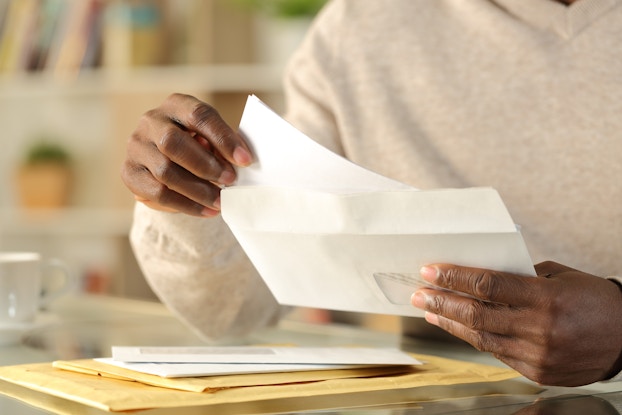 Close-up on a pair of hands opening a letter. A small pile of envelopes and manila folders sits on a table beneath the hands.
