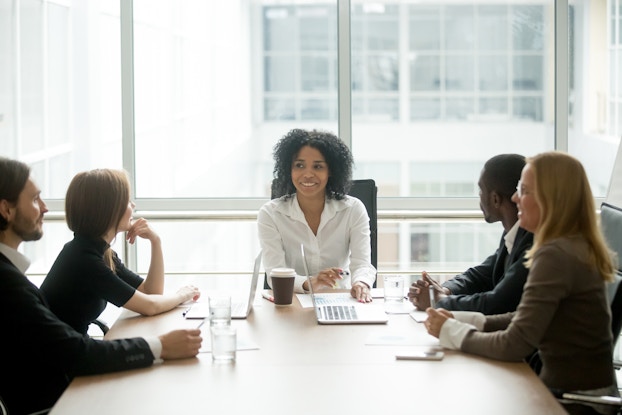  A diverse group -- three woman and two men of various races -- sits around a conference room table. At the head of the table is a smiling Black woman; behind her is a floor-to-ceiling window..