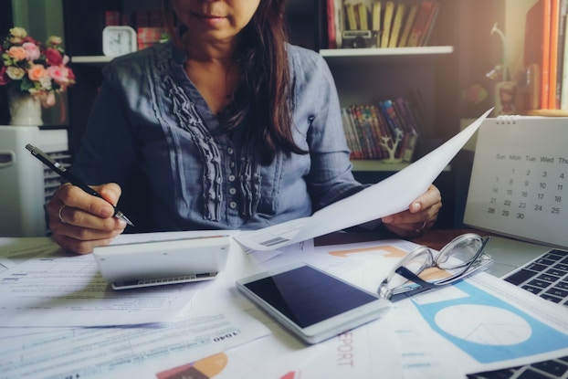 A woman, her face partially cut off in the photo, sits at a desk scattered with papers, a smartphone, and a laptop. She holds a paper and pencil and consults a calculator in front of her.