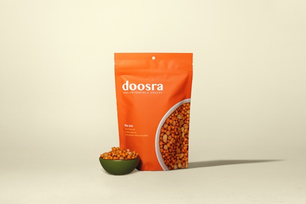 An orange bag of doosra's Indian-inspired snacks next to a small green bowl of the snacks -- a mix of boondi (chickpea puffs), roasted peanuts, and caramelized white chocolate, coated with a masala spice blend that adds an orange tint to the mix.