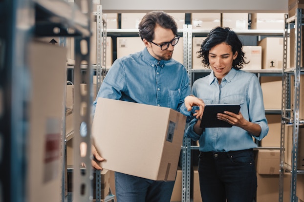 A man and a woman stand next to each other in an aisle in a warehouse. The man wears a chambray shirt and dark-rimmed glasses and holds a cardboard box. The woman wears a lighter blue chambray shirt and holds an electronic tablet. The man uses one finger to point at something on the tablet's screen. The metal shelves around the two people are filled with more cardboard boxes.