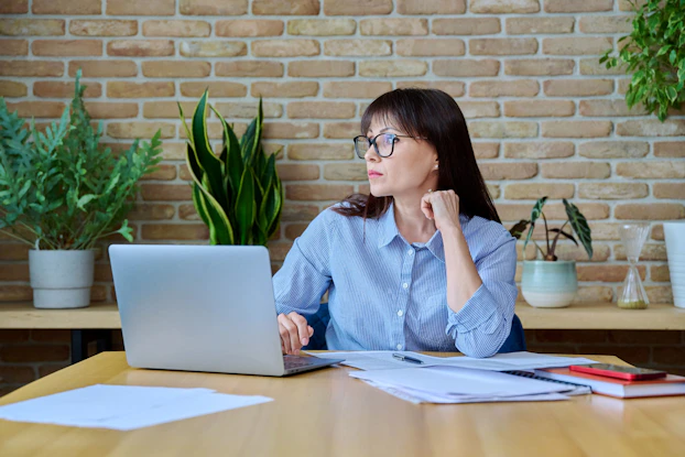 A woman is seated at a desk in front of a laptop. Her head is turned to the side and she is deep in thought. She has papers spread out in front of her.