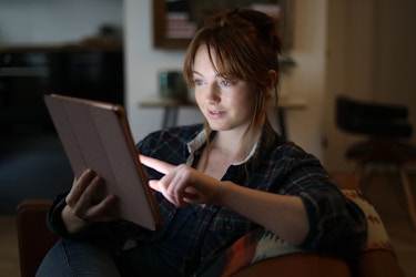  A young woman is sitting in her home. She is holding a tablet and perusing online goods. Her face is illuminated by the glow of the tablet. 