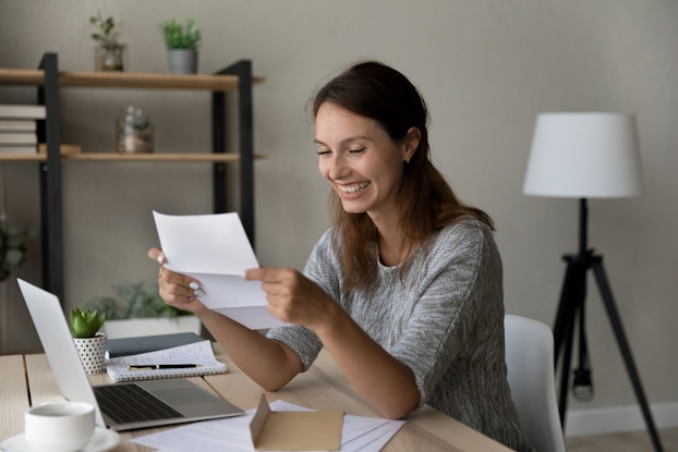 A young woman sits at a table and looks at an unfolded paper in her hands with a big smile on her face. The table in front of her holds an open laptop, an open envelope, several other papers, and a notebook and pen. The room in the background has neutral gray walls; behind the woman are a three-legged lamp and a shelf holding several potted plants.
