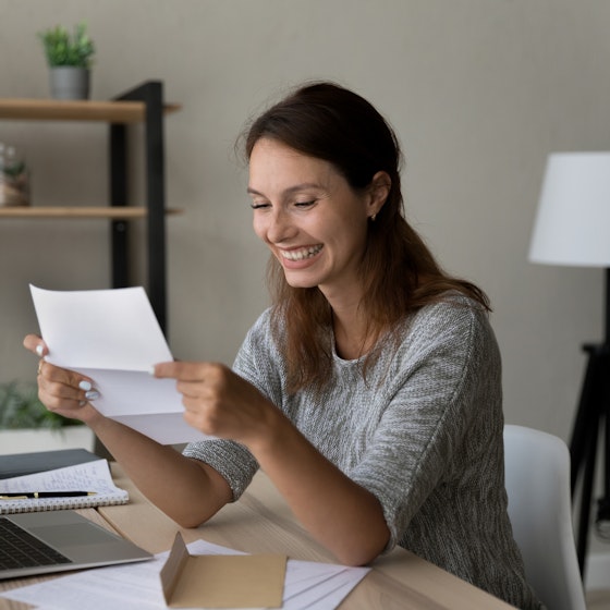 A young woman sits at a table and looks at an unfolded paper in her hands with a big smile on her face. The table in front of her holds an open laptop, an open envelope, several other papers, and a notebook and pen. The room in the background has neutral gray walls; behind the woman are a three-legged lamp and a shelf holding several potted plants.