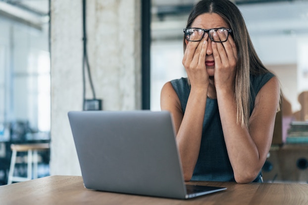 A long-haired woman sits at a table in front of an open laptop with her hands covering her eyes, looking exhausted. She's wearing glasses, which sit at the end of her fingertips, pushed out of the way by her hands.