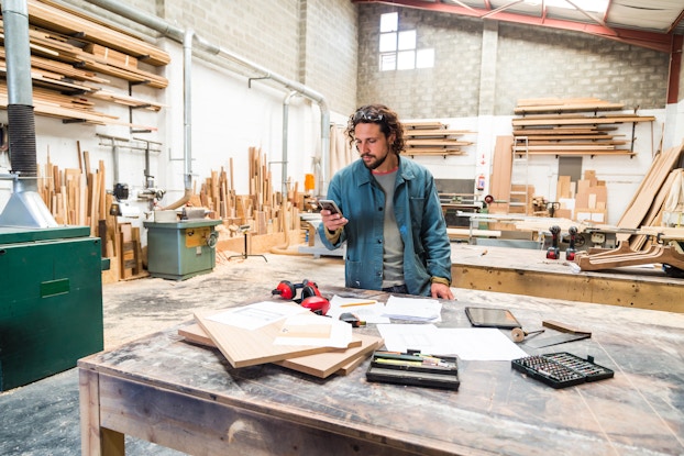 A wide shot of a man behind of a large wooden worktable in a cavernous warehouse. The man is looking down at the smartphone in his right hand. He has a beard and curly dark blond hair, and he wears a chambray jacket over a light gray T-shirt. Safety goggles are pushed up on top of his head. In the background, planks of wood of various lengths are standing against the walls of the warehouse and more planks are stacked on shelves higher up.