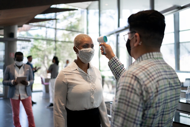  A man holds a digital thermometer to a woman's forehead. Another man stands several feet behind the woman, looking at a digital tablet. Everyone in the picture is wearing a face mask.