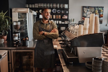  A young man stands behind the service counter at a cafe. He leans against the edge of the counter with his arms folded and a confident smile on his face. The man wears a dark gray pinstriped apron over an olive green long-sleeved polo shirt. On top of the part of the counter running parallel to the man is a large coffee machine, on top of which are piles of cups and mugs. 