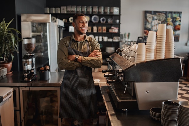  A young man stands behind the service counter at a cafe. He leans against the edge of the counter with his arms folded and a confident smile on his face. The man wears a dark gray pinstriped apron over an olive green long-sleeved polo shirt. On top of the part of the counter running parallel to the man is a large coffee machine, on top of which are piles of cups and mugs.