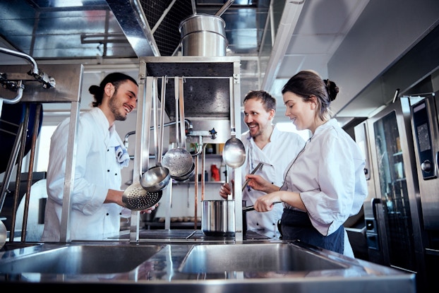 Three chefs stand around a chrome stovetop, smiling and laughing. They all wear white chef's jackets, and the chef on the far right, a woman with dark brown hair pulled back into a bun, is stirring something in a large silver pot.