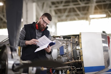  A man stands in an aircraft hanger next to a cylindrical airplane engine and reads a book. The man is bearded and wears a black vest over a gray plaid shirt. He also wears safety goggles and, around his neck, a pair of orange headphones. The airplane engine has part of its cover removed, showing the complex mechanisms inside. 