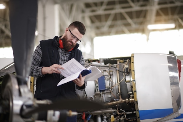  A man stands in an aircraft hanger next to a cylindrical airplane engine and reads a book. The man is bearded and wears a black vest over a gray plaid shirt. He also wears safety goggles and, around his neck, a pair of orange headphones. The airplane engine has part of its cover removed, showing the complex mechanisms inside.