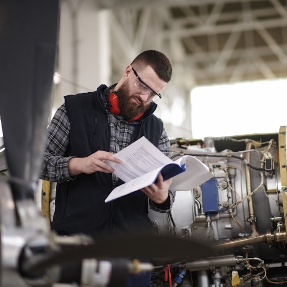 A man stands in an aircraft hanger next to a cylindrical airplane engine and reads a book. The man is bearded and wears a black vest over a gray plaid shirt. He also wears safety goggles and, around his neck, a pair of orange headphones. The airplane engine has part of its cover removed, showing the complex mechanisms inside.