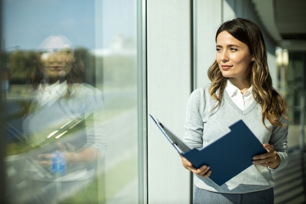 A woman stands next to a floor-to-ceiling window, holding an open blue binder. The woman has wavy brown hair with gold highlights and wears a light gray sweater over a white collared shirt. The window reflects the woman's image back at her.