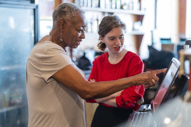  Two women, one older and one teenaged, stand at a computer at a counter in a cafe or restaurant. The older woman has short salt-and-pepper hair and is wearing a pale gray T-shirt; she stands in profile to the viewer and is pointing at the screen of the computer monitor. The teenager wears a red shirt with loose sleeves; she stands facing the viewer with her arms folded and her head turned to look at the screen of the computer.In the background is a refrigerator with a glass-fronted door fogged with condensation and several shelves mounted on a back wall.